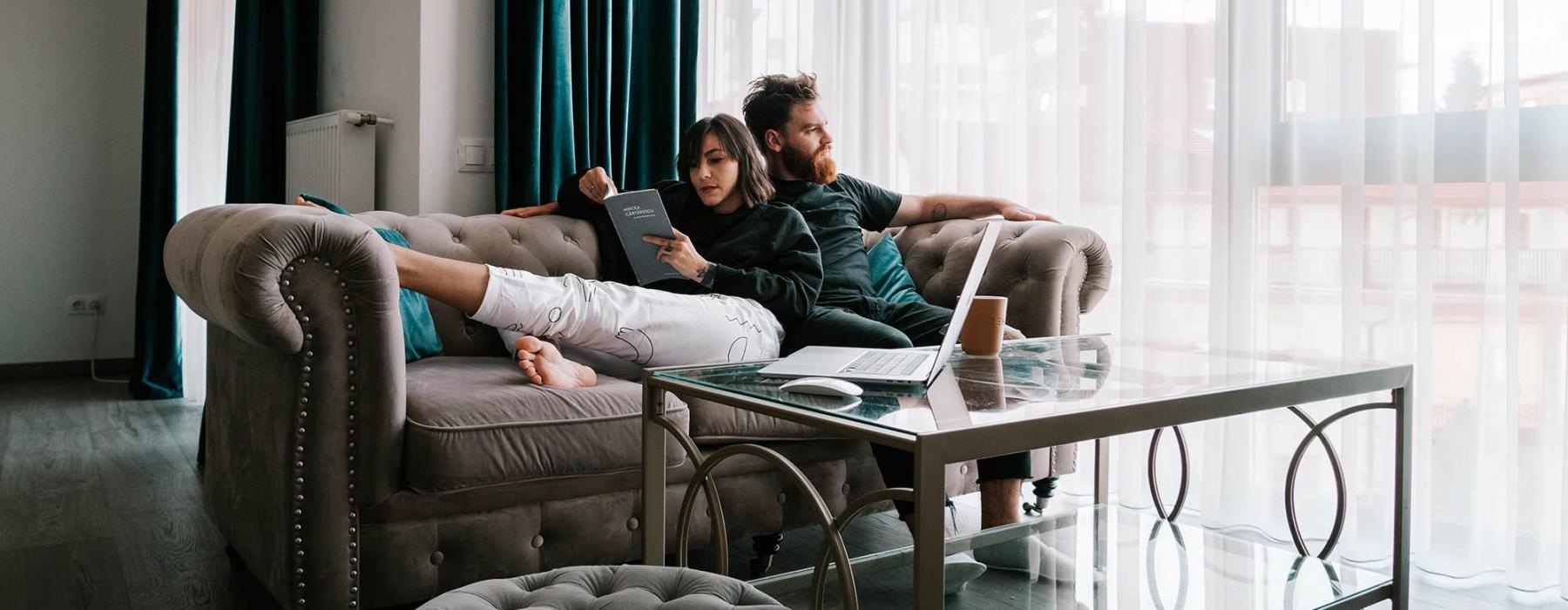 man sits on a couch and looks out the window while his wife lies against him reading a book