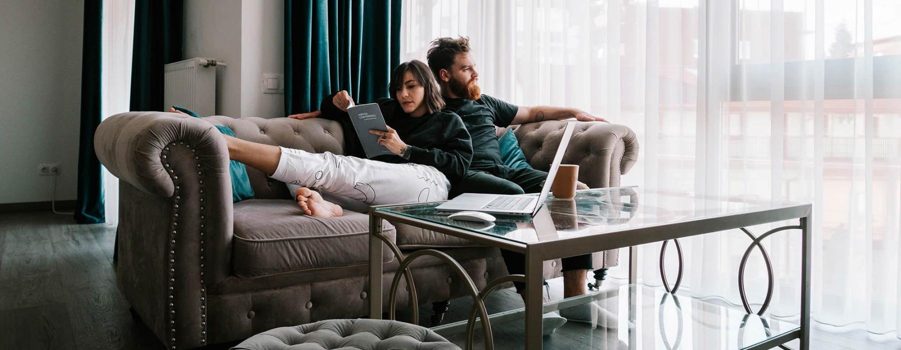 man sits on a couch and looks out the window while his wife lies against him reading a book